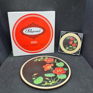 May include: A round Pimpernel acrylic casserole stand featuring a black surface with a floral design of red nasturtium flowers and green leaves. The set includes a square white box with red circular accents and a smaller matching round coaster.
