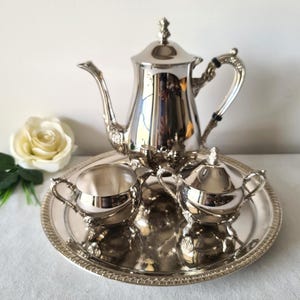 May include: A silver tea set with intricate detailing, comprising a teapot, creamer, sugar bowl, and a small dish, presented on a round tray. The set is highly reflective, with floral accents on the handle and lid.