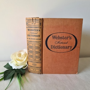 Peut inclure: Deux dictionnaires nationaux Webster's vintage. L'un est debout, montrant le dos avec du texte et des éléments décoratifs. L'autre est à plat, avec le titre dans un ovale. Une rose blanche avec des feuilles vertes est au premier plan.