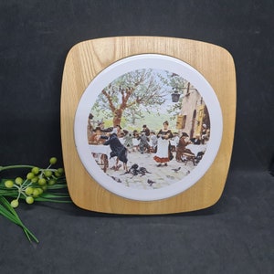 Könnte beinhalten: Dekorative Holzplakette mit einem runden, gerahmten Druck einer europäischen Freiluftcafé-Szene. Der Druck zeigt Menschen beim Essen, mit einer Frau in weißer Schürze und rotem Rock. Der Holzrahmen hat eine hellbraune Oberfläche.