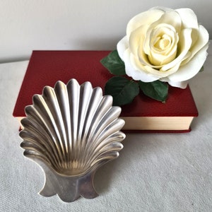 May include: A silver-coloured, shell-shaped dish with a fluted design. A white rose with green leaves rests on a red book. The dish is on a light grey surface.