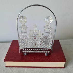 Może przedstawiać: Zestaw toaletowy z kryształu w stylu vintage z dwiema butelkami i małym kwadratowym pojemnikiem, wszystkie z fasetowanymi korkami. Zestaw jest trzymany w srebrnym metalowym uchwycie i stoi na czerwonej książce.