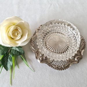 Pode incluir: Um prato de vidro transparente com um padrão de diamante está sobre um prato prateado e ornamentado. Uma rosa branca e folhas verdes estão à esquerda do prato. O prato e o prato estão sobre uma superfície branca.