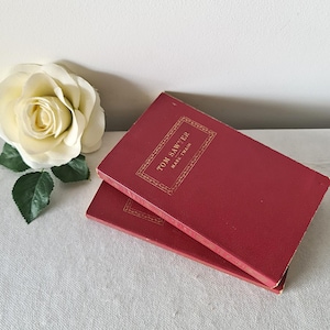 Op de afbeelding: Twee gebonden boeken getiteld Tom Sawyer van Mark Twain met dieprode kaften en goudkleurige letters in reliëf binnen een decoratieve rechthoekige rand, vergezeld door een grote crèmekleurige kunstmatige roos met groene bladeren.