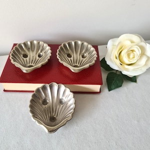May include: Three silver seashell-shaped dishes. They are arranged on a red book, with a white rose in the background. The dishes have a ribbed texture and two small holes.
