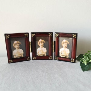 May include: A triptych photo frame with a dark red wooden finish. Each frame holds a vintage portrait of a young girl. The frames are connected by brass hinges and adorned with gold-coloured floral accents. The overall design is classic and elegant.