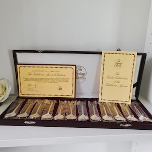 Könnte beinhalten: Eine Sammlung von Silberlöffeln in einer dunklen Holzkiste. Die Löffel sind einzeln verpackt und ordentlich angeordnet. Ein Echtheitszertifikat und ein Büchlein mit dem Titel "The Twelve Celebrities of the Tichborne Spoons" begleiten die Löffel.