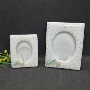 May include: Two white ceramic photo frames with oval apertures. They feature a floral pattern of pink roses and green leaves. A blue ribbon with pink roses adorns the bottom of each frame.