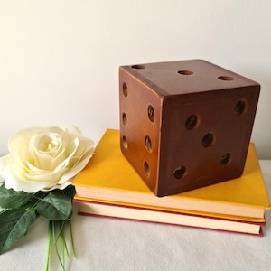 Könnte beinhalten: Ein großer, brauner Würfel aus Holz mit gebohrten Punktmarkierungen, der auf zwei Büchern mit gelben und roten Einbänden liegt. Eine weiße Rose und grüne Stiele befinden sich im Vordergrund. Der Würfel ist ein dekorativer Gegenstand.