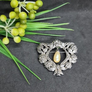 May include: A silver brooch in a heart shape, decorated with floral patterns and a bow. A cream-coloured, pear-shaped pearl hangs from the centre. Green sprigs and yellow berries are in the background.