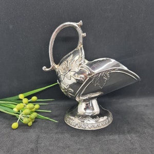 May include: Silver-plated sugar bowl with a handle and floral detailing. The bowl has a curved shape and sits on a pedestal base. The handle is arched and has decorative elements. The design includes roses and leaf patterns.