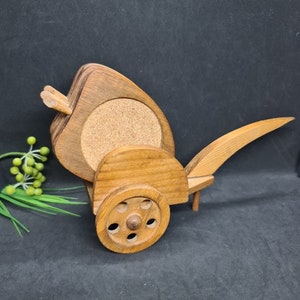 May include: A wooden cart-shaped holder containing several round coasters with cork centres. The holder features a natural wood grain finish, a single wheel on the side, and a long handle extending from the back, designed to store and display coasters.