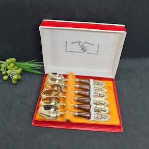 Könnte beinhalten: Ein Satz Vintage-Löffel in einer roten Schachtel mit weißem Deckel. Die Löffel haben goldfarbene Schalen und verzierte Griffe mit brauner Holzmaserung. Die Schachtel ist mit rotem Samt und gelbem Stoff ausgekleidet.