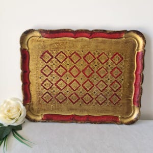 Puede incluir: Bandeja decorativa rectangular con superficie dorada y borde rojo. La bandeja presenta un patrón repetitivo de diamantes rojos y detalles florales dorados. Los bordes son festoneados.