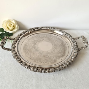 May include: A silver-plated serving tray with a detailed grape and vine border and ornate handles. The tray has an oval shape and a central embossed floral pattern. The handles are intricately designed. A white rose is visible in the background, adding to the presentation.