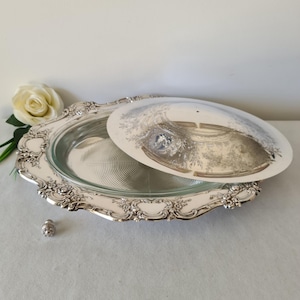 Könnte beinhalten: Eine silberbeschichtete Servierschale mit Glaseinsatz und dekorativem Deckel. Die Schale hat ein verziertes, florales Design am Rand. Ein kleines, dekoratives Silberobjekt befindet sich in der Nähe der Schale. Eine weiße Rose ist im Hintergrund.