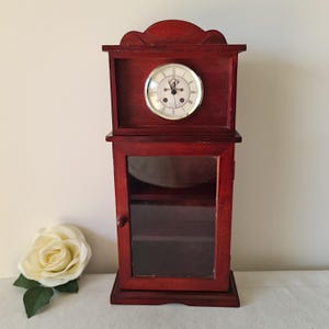 May include: A miniature, dark red wooden clock with a glass cabinet. The clock face is white with black Roman numerals and hands. A white rose is next to the clock.