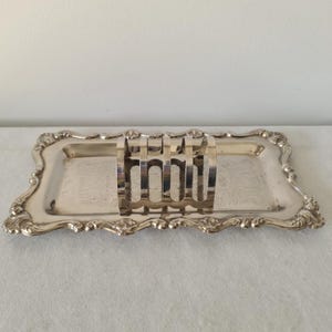 May include: Silver-plated toast rack and serving tray. The rectangular tray has ornate edges and a floral design. The toast rack has six slots. The set is made of silver-colored metal and is likely vintage.