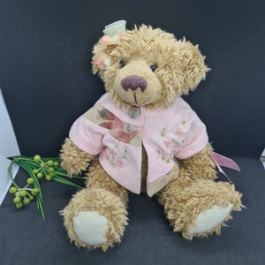 Op de afbeelding: Een lichtbruine teddybeer met een roze jasje met bloemenborduurwerk en patches. De beer heeft een roze strik en bloem op zijn hoofd. De beer zit op een zwarte ondergrond.