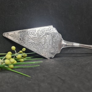 Puede incluir: Servidor de pastel plateado con un diseño floral y de hojas ornamentado. El servidor tiene una hoja triangular con un borde dentado y un mango largo. El patrón floral incluye flores y hojas, y un emblema central.