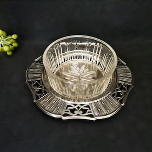 May include: A clear glass bowl with a starburst design sits atop an ornate silver-colored metal dish. The dish has a decorative, openwork edge and a circular base. The background is a solid black.