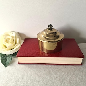 Può includere: Un piccolo contenitore antico in ottone con coperchio a gradini è appoggiato su un libro rosso. Il contenitore ha un pomello decorativo in cima. Una rosa bianca è sullo sfondo. Il contenitore è probabilmente usato per riporre piccoli oggetti.