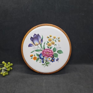 Könnte beinhalten: Runder, weißer Keramik-Topfuntersetzer mit floralem Design. Das Design zeigt eine lila Iris, eine rosa Pfingstrose, gelbe und blaue Blumen und grüne Blätter. Der Untersetzer hat einen Holzrahmen.