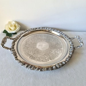 Pode incluir: Uma bandeja redonda prateada com detalhes ornamentados. A bandeja apresenta uma borda e alças decorativas, e um padrão floral gravado no centro. Uma rosa branca está no fundo.