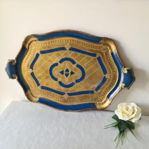 以下が含まれることがあります： 金色の表面と青い縁取りが施された、装飾的な長方形のトレイ。トレイは、中央に四つ葉のクローバーの形をした幾何学模様が特徴です。トレイの両側にハンドルがあり、長さは約51cmです。