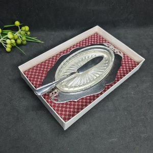 Op de afbeelding: Een vintage boterservies. Het bevat een heldere, ovale glazen schaal met een geribbeld ontwerp, een zilverkleurig mes en een decoratief zilverkleurig dienblad. De set wordt gepresenteerd in een witte doos met een rood patroon.