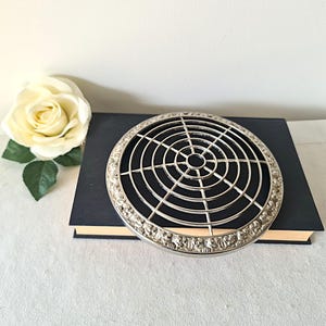 May include: A silver flower frog with a circular grid design, placed on a dark blue book. The flower arranger has a decorative edge. A cream rose is in the background. The flower frog is approximately 15 cm in diameter.