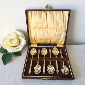 Könnte beinhalten: Set mit sechs Silberlöffeln in einem braunen Kunstleder-Etui mit goldenem Verschluss. Die Löffel haben verzierte Griffe und ovale Laffen. Das Etui hat ein braunes Samtinterieur und ein goldenes Satinfutter. Eine weiße Rose ist im Hintergrund.