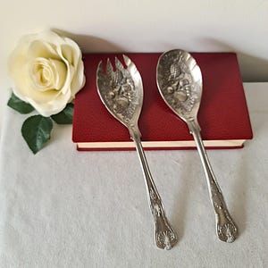 Op de afbeelding: Een paar sierlijke zilveren serveerbestekken, een lepel en een vork, met gedetailleerde fruit- en bloemmotieven op de koppen. De bestekken liggen op een rood boek, met een witte roos op de achtergrond.