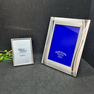Könnte beinhalten: Zwei silberbeschichtete Fotorahmen. Der größere Rahmen hat ein wellenförmiges Design und einen blauen Hintergrund. Der kleinere Rahmen hat eine rechteckige Form. Beide Rahmen tragen den Text "Silver Plated" und "Bowon". Der größere Rahmen ist 9 x 13 cm groß.