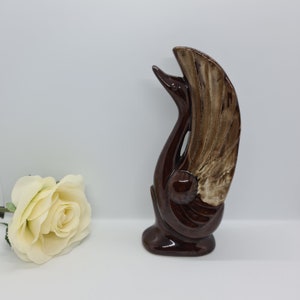 May include: A ceramic swan figurine featuring a glossy dark brown finish on the body and neck, with a textured, lighter tan and cream-coloured wing detail. The swan is depicted with its head tilted upward and its wing extended vertically.