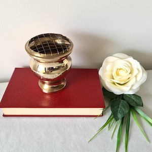May include: A brass flower arranger with a grid top, placed on a red book, beside a white rose. The flower arranger has a round base and a bulbous body. The rose has a yellow centre and green leaves.