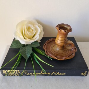 May include: A brown ceramic candle holder with a ruffled top and a circular base. A white rose and green grass are next to the candle holder on a black book with gold lettering that reads "Roberta Atow Cannonberry Chase".