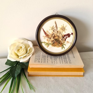 Op de afbeelding: Een ronde, ingelijste bloemstuk met gedroogde bloemen in bruin, beige en rood. De compositie staat tegen een witte achtergrond en is omlijst in een donkerbruine lijst. De lijst staat op twee gele boeken.