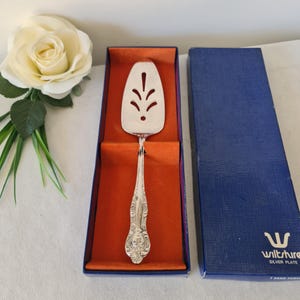Peut inclure: Une pelle à gâteau argentée avec un manche décoratif, présentée dans une boîte bleue avec un intérieur orange. La pelle présente une lame ajourée avec un motif floral. La boîte porte le texte "Wiltshire Silver Plate".