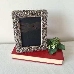 Pode incluir: Uma moldura retangular prateada com volutas ornamentais está em cima de um livro vermelho. A moldura tem um interior preto e é decorada com padrões detalhados e giratórios. Um pequeno ramo de vegetação repousa ao lado da moldura.