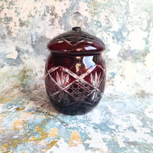 Op de afbeelding: Een robijnrode, geslepen glazen pot met een deksel en een heldere, ronde knop. De pot heeft een gedetailleerd, geometrisch patroon met een diamant- en bladerontwerp. De pot wordt waarschijnlijk gebruikt voor opslag of weergave.