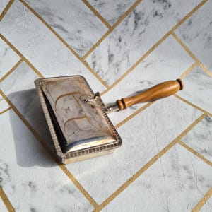 Peut inclure: Un ramasse-miettes argenté vintage avec une poignée en bois. Le plateau rectangulaire a un bord décoratif et un couvercle à charnière. La poignée est de couleur marron clair. Le ramasse-miettes est conçu pour ramasser les miettes sur une table.