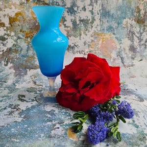 May include: A light blue glass vase with a flared top and a stemmed base. A large red rose and a sprig of small purple flowers are next to the vase. The background has a distressed, textured appearance with shades of blue, white, and brown.