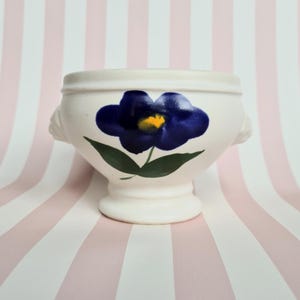 Peut inclure: Bol en céramique crème avec un motif de fleur bleue. La fleur a un centre jaune et des feuilles vertes. Le bol a deux petites poignées et un socle. Le fond est un motif à rayures roses et blanches.