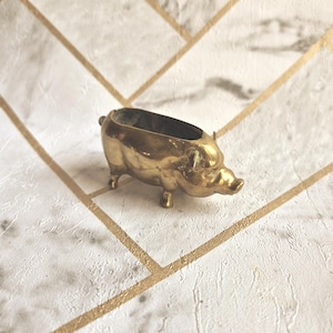 Könnte beinhalten: Eine kleine, hohle Messingfigur in Form eines Schweins. Das Objekt hat eine glatte, goldfarbene Metalloberfläche mit poliertem Finish, vier kurze Beine und eine offene Oberseite, die eine gefäßartige Struktur mit Schnauze und Ohren bildet.
