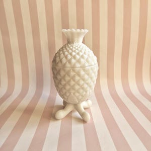 Peut inclure: Un pot à couvercle en verre opalin blanc en forme d'ananas avec un motif en losange texturé sur le corps et le couvercle. Le pot présente un bord festonné sur le dessus et repose sur trois pieds arrondis et incurvés à la base, créant une structure surélevée.