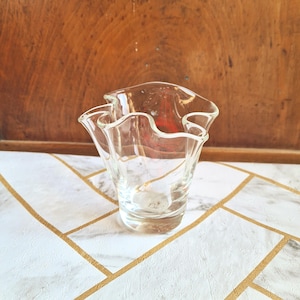 Pode incluir: Um vaso de vidro transparente com uma borda ondulada. O vaso tem uma abertura larga e uma base curta e arredondada. O vidro é transparente, permitindo que a luz passe. O vaso é provavelmente projetado para exibir flores ou outros itens decorativos.
