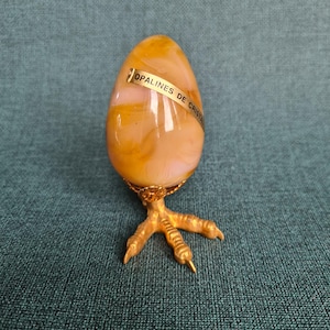 Könnte beinhalten: Ein eiförmiges Deko-Objekt mit einem goldfarbenen Metallsockel in Form eines Vogelfußes. Das Ei hat gelbe und rosa Farbtöne, mit einem goldenen Band mit der Aufschrift "OPALINES DE CRISTAL".
