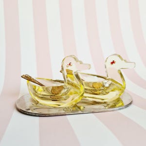 Op de afbeelding: Twee heldere glazen eendvormige bakjes met geel getinte lichamen en roze snavels, rustend op een zilverkleurige ovale schaal. Eén eend heeft een klein lepeltje. De achtergrond heeft roze en witte strepen.