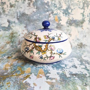 Könnte beinhalten: Keramik-Schmuckdose mit Deckel, verziert mit einem Blumenmuster mit rosa, blauen und gelben Blüten auf einem geknackten weißen Hintergrund. Dunkelblauer Rand und passender Knopf auf dem Deckel.